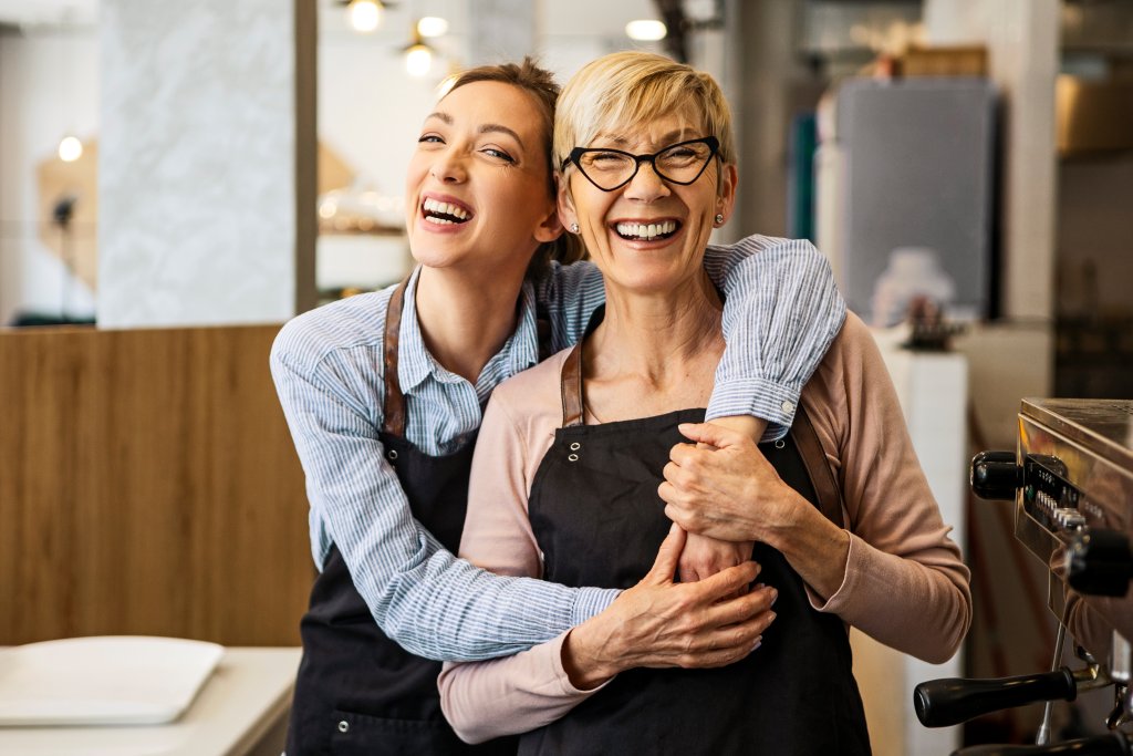 Zwei lachende Frauen in einer Caféküche, eine trägt eine schwarze Schürze, beide umarmen sich.