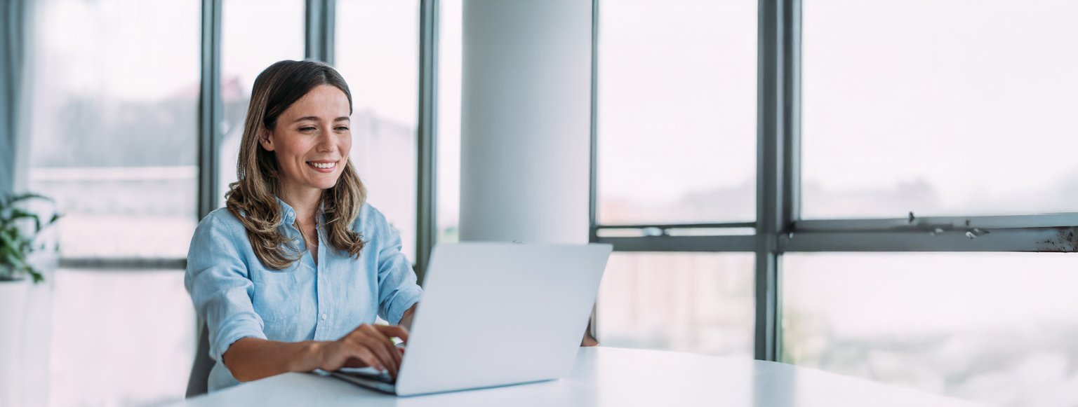 Frau arbeitet an einem Laptop in hellem Büro mit Fensterblick und Pflanzen.