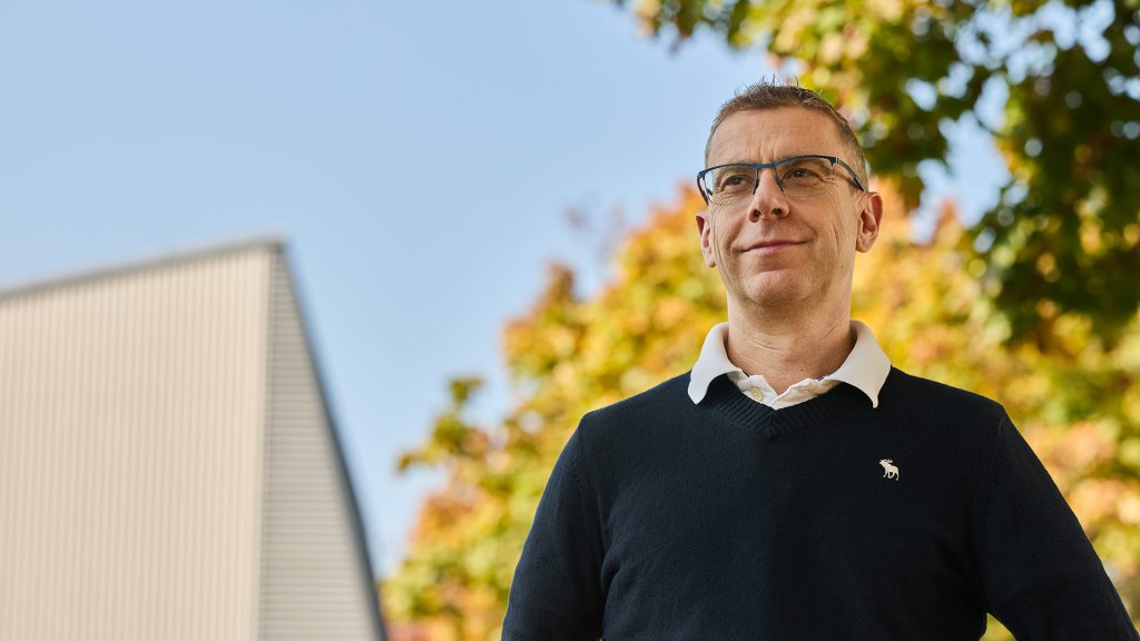 Michael Steiner steht im Freien vor einem modernen Gebäude mit bunten Herbstbäumen im Hintergrund.