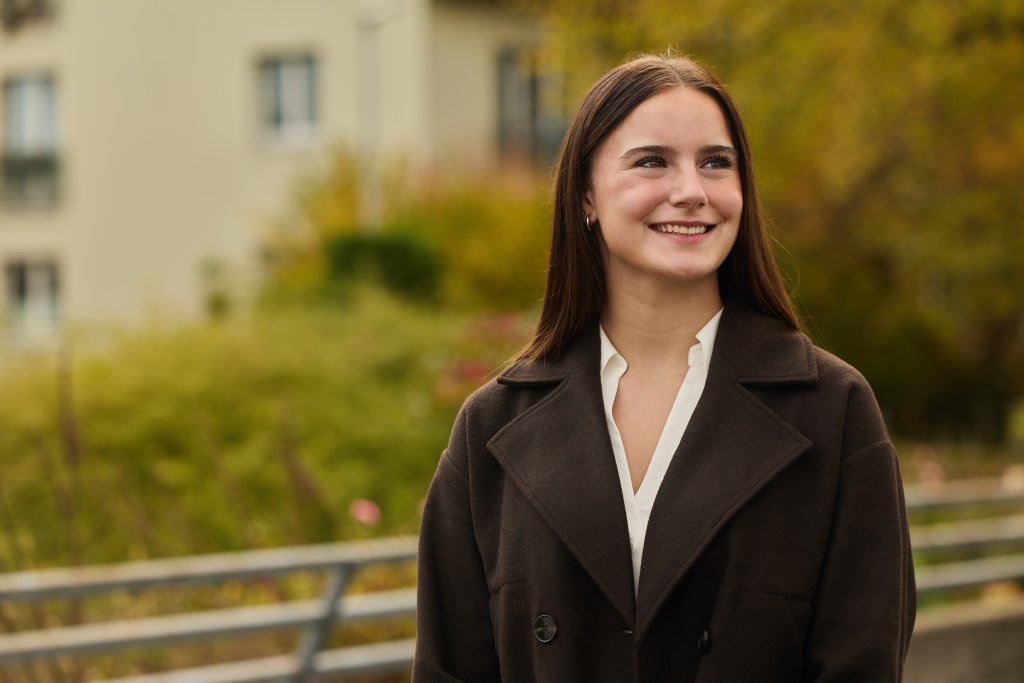Sienna Gschwind in dunklem Mantel vor Naturhintergrund bei Tageslicht.