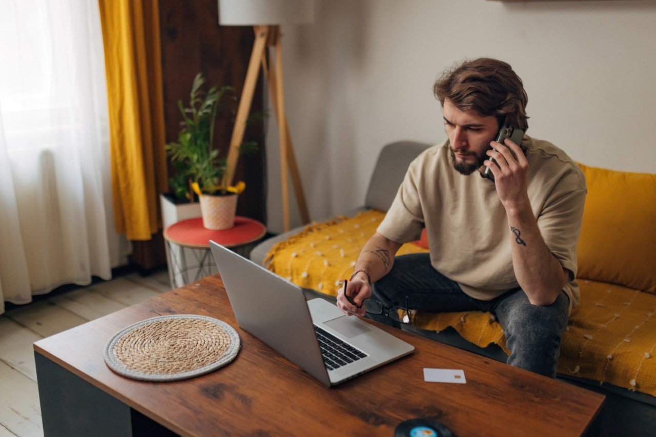 Ein Mann mit Bart und braunen Haaren sitzt auf seinem Sofa und schaut an seinem Tisch seinen Laptop an, während er am telefonieren ist und in seiner rechten Hand eine Brille hat. aus dem Artikel Wie kann ich mich effektiv vor Online-Betrügern schützen?