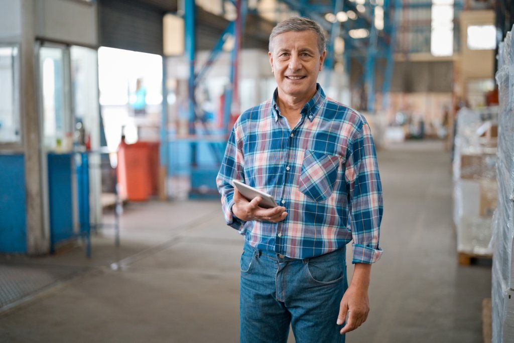 Mann im Blaumann steht in einer Fabrikhalle und hält ein Tablet in der Hand.