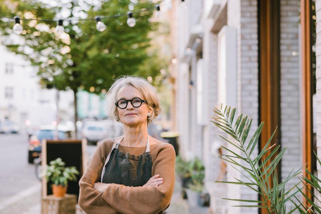 Porträt einer selbstbewussten älteren Floristin, die vor ihrem Laden steht. Reife Frau mit Schürze, verschränkten Armen und Blick in die Kamera, während sie vor ihrem Blumenladen steht.