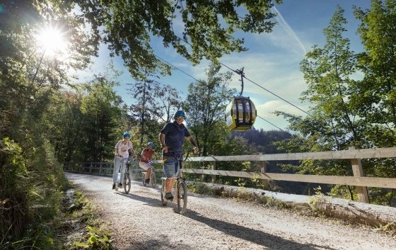 Drei Personen fahren mit ihren Fahrrädern durch den Wald. Neben ihnen befindet sich eine Luftseilbahn und eine Gondel fährt vorbei.