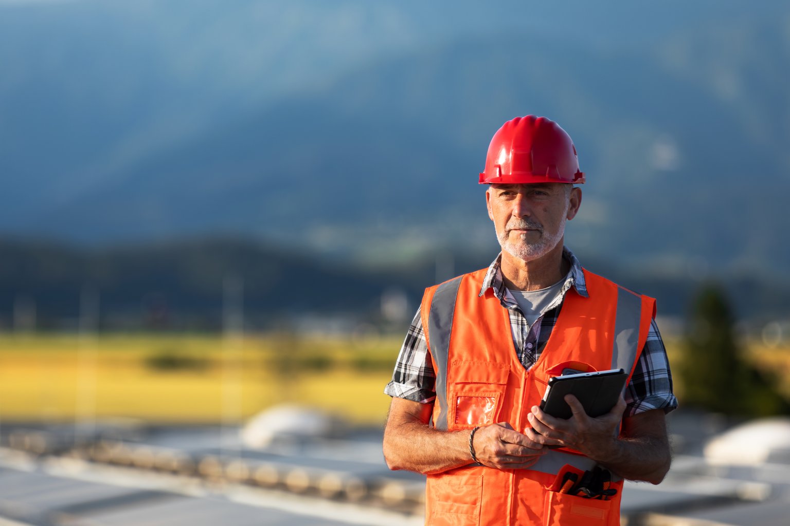 Ein Bauarbeiter in einer orangefarbenen Weste und einem Sicherheitshorn hält ein Tablet im Freien.