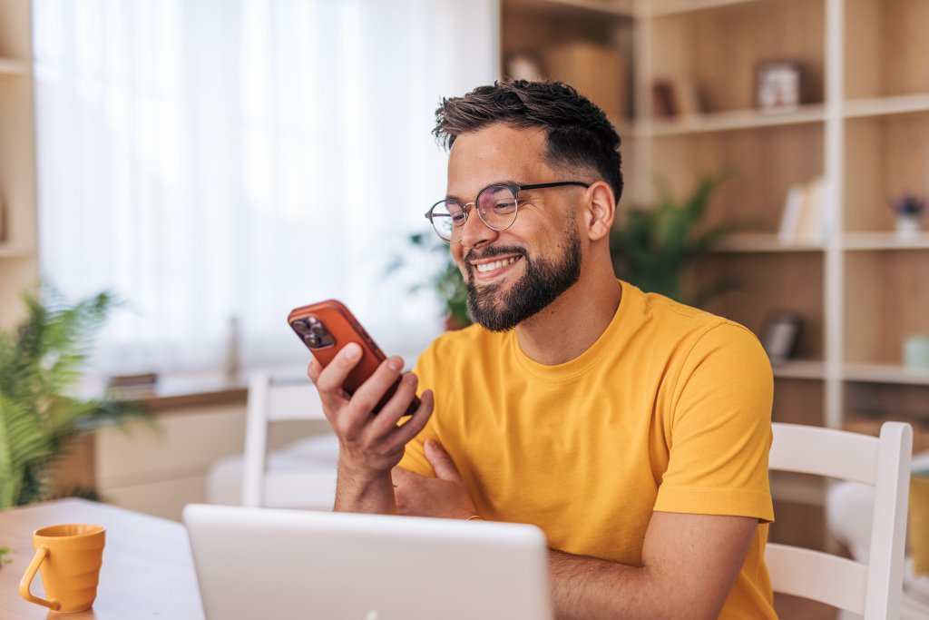 Junger Mann sitzt am Schreibtisch zu Hause, lächelt beim Blick auf sein Smartphone, Laptop davor