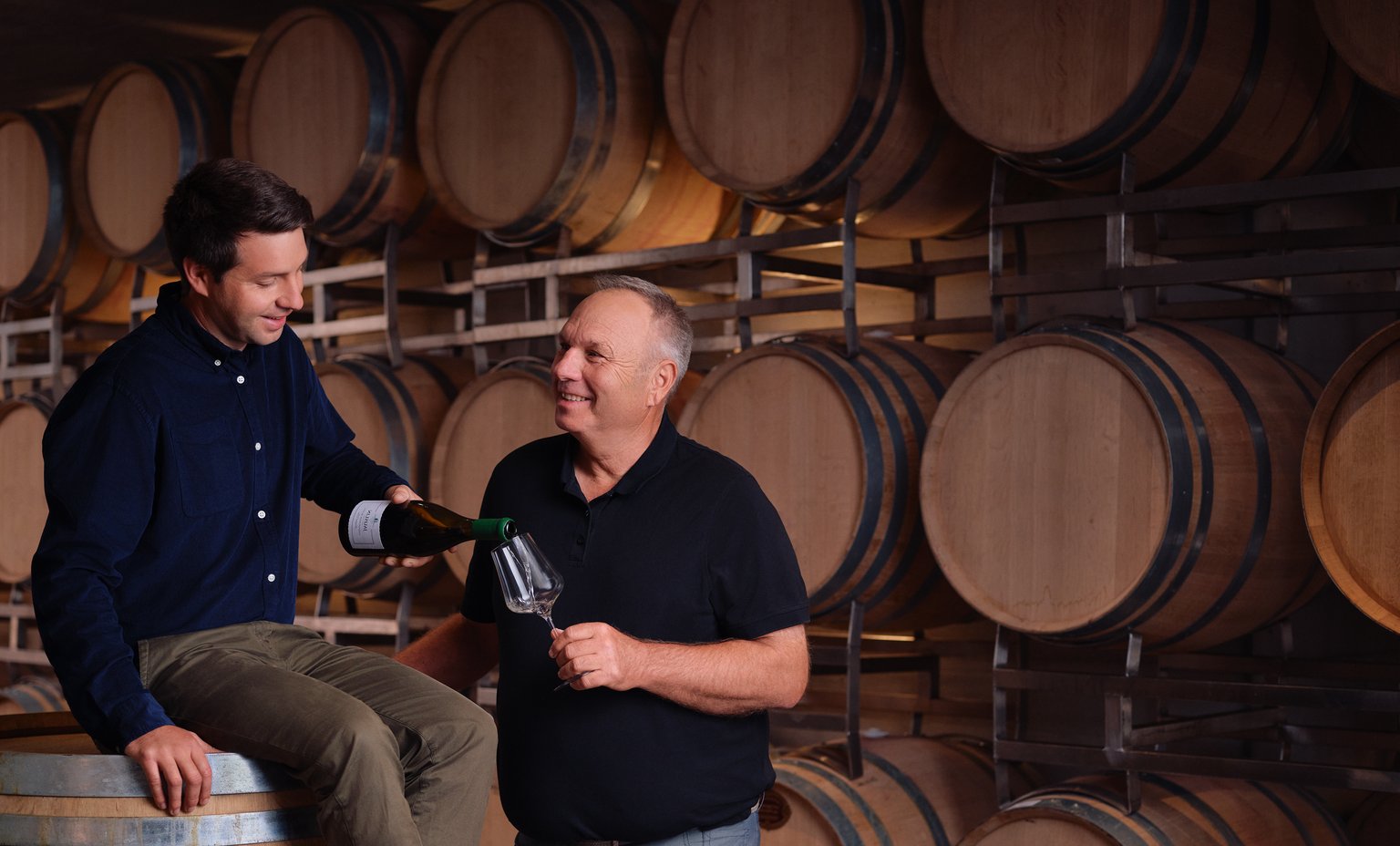 Zwei Männer im Weinkeller, einer schenkt dem anderen Wein ein.