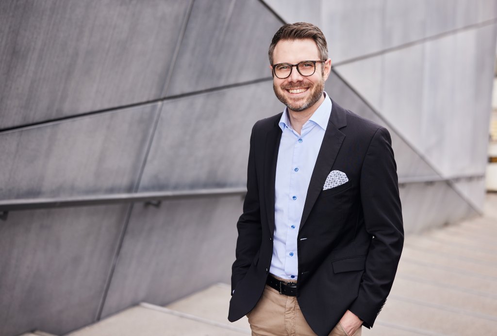 Ein freundlicher Mann im Anzug posiert auf einer modernen Treppe vor einer geometrischen Wand.