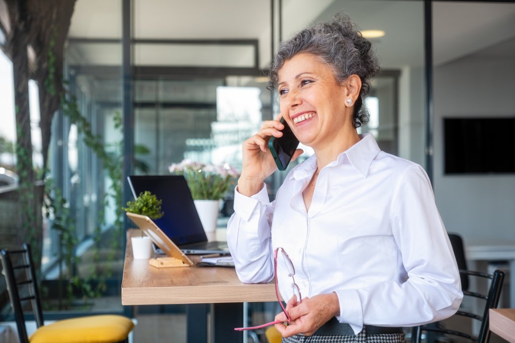 Frau mit Brille telefoniert, steht in einem modernen Büro mit Laptop und Pflanzen.