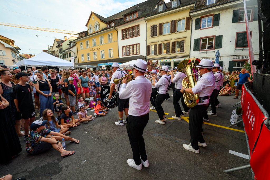 Eine Blasmusikkapelle spielt auf der Straße, Zuschauer beobachten die Aufführung.