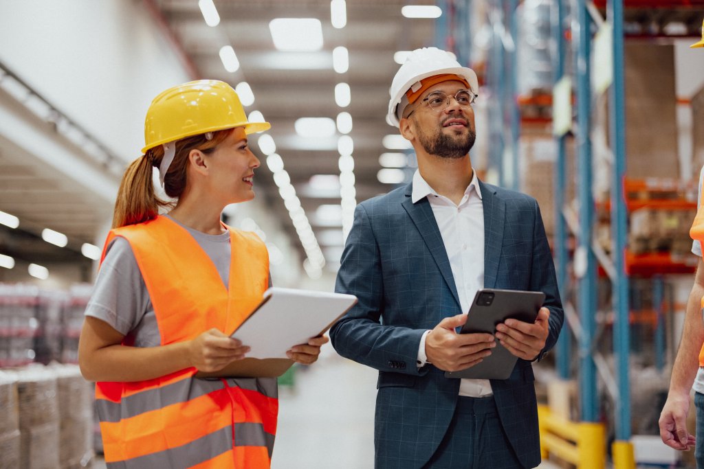 Zwei Fachleute diskutieren in einem Lagerhaus, die Frau in Schutzkleidung, der Mann im Anzug.