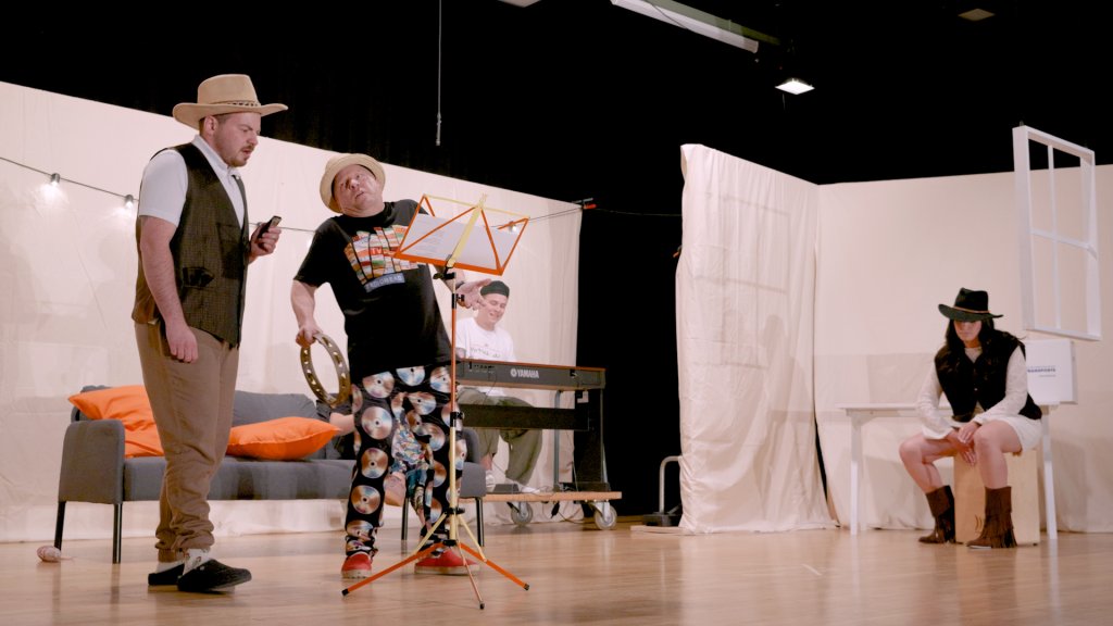 Drei Theaterakteure auf Bühne, eine trägt Hut, andere sitzen oder spielen Klavier