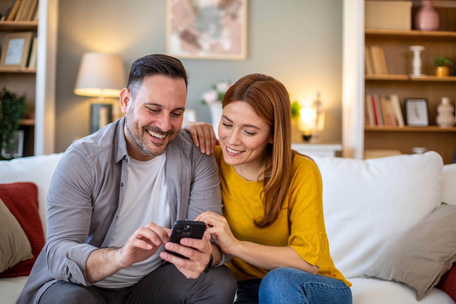 Zwei Personen sitzen auf einer Couch, schauen auf ein Smartphone, im Wohnzimmer. aus dem Artikel Vermögen aufbauen leicht gemacht
