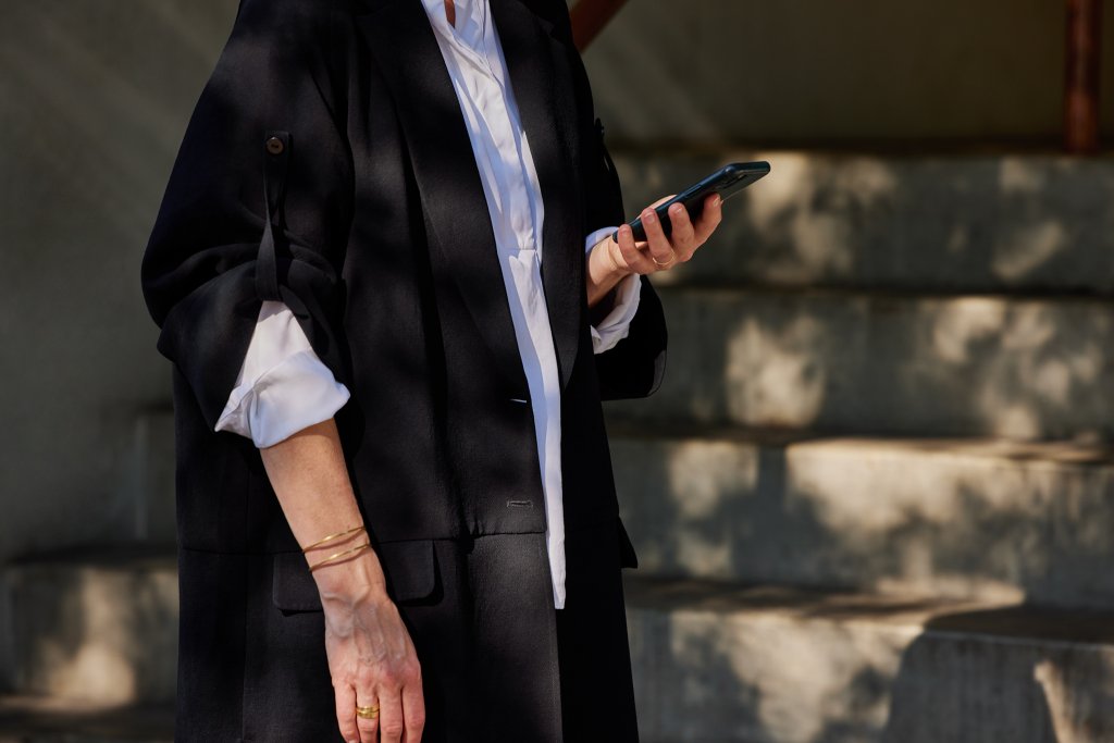 Eine junge Frau in einem schwarzen Mantel hält ihr Smartphone in der Hand.