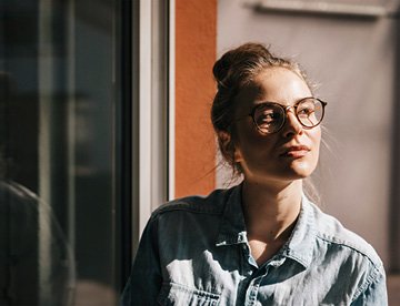 Frau mit Brille schaut nachdenklich aus dem Fenster in einem hellen Raum. aus dem Artikel Frauenpower beim Anlegen
