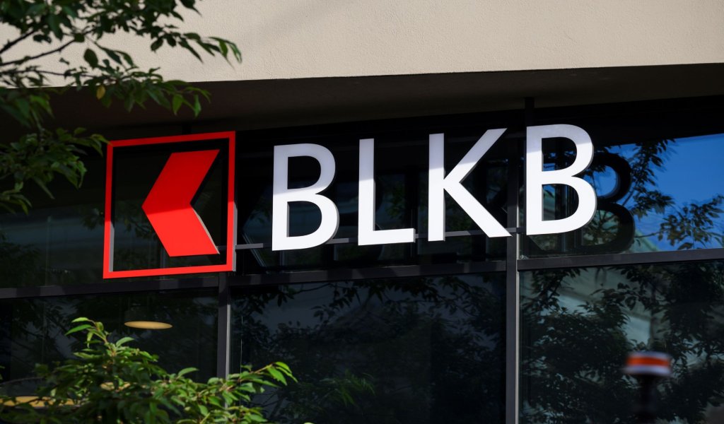 Schriftzug der Basellandschalftlichen Kantonalbank (BLKB) mit rotem Logo an einer Gebäudefront, reflektierende Fenster und Grünpflanzen. aus dem Artikel