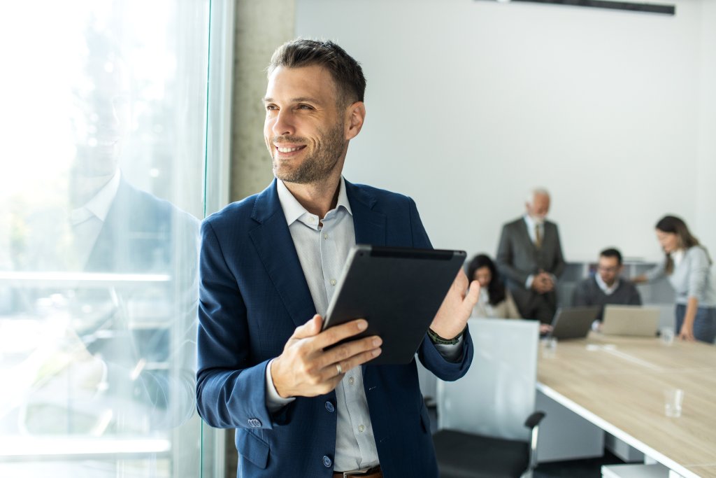 Lächelnder Geschäftsmann, der ein Tablet in einem modernen Büroumfeld mit Kollegen im Hintergrund hält.