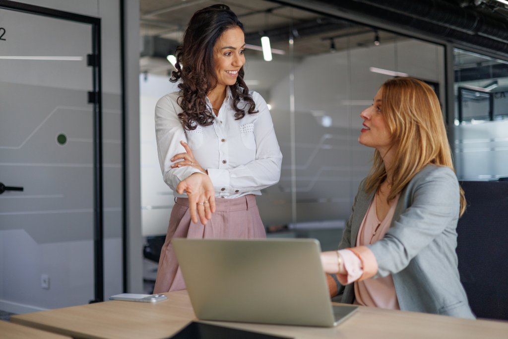 Zwei Frauen im Büro, arbeitet an Laptop, unterhalten sich, im Hintergrund Glasraum