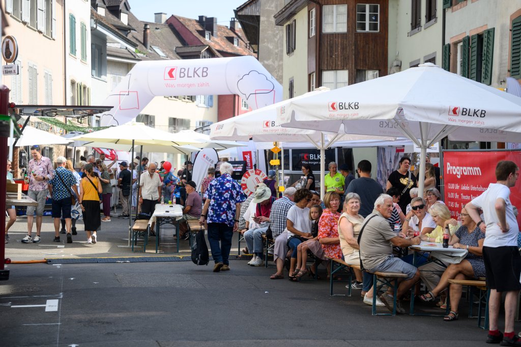 Menschen sitzen und stehen an Tischen und Schirmen in einer belebten Straße.