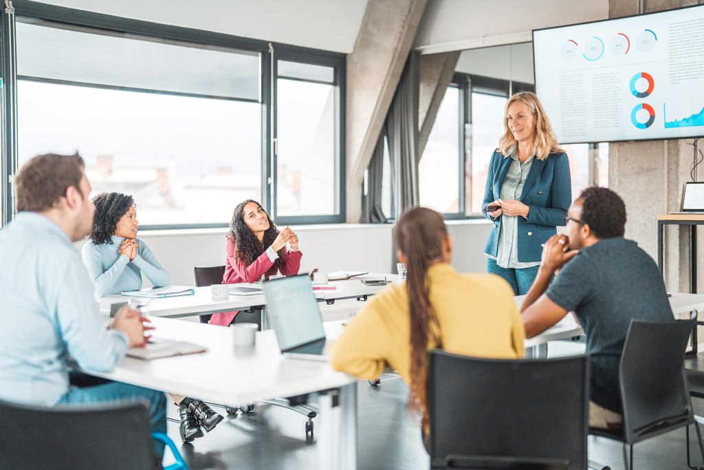 Unternehmerinnen und Unternehmer sitzen in einem Raum und hören einer Frau mittleren Alters zu die Statistiken des Unternehmens auf einem grossen Bildschirm zeigt