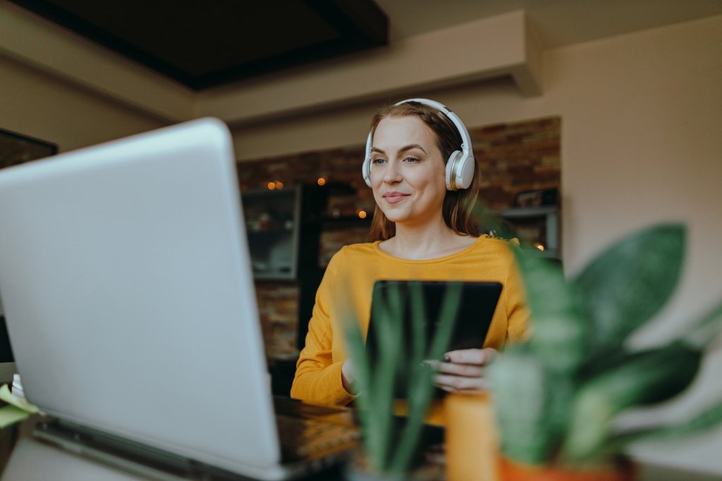 Junge Frau mit Kopfhörer und Tablet arbeitet am Laptop.