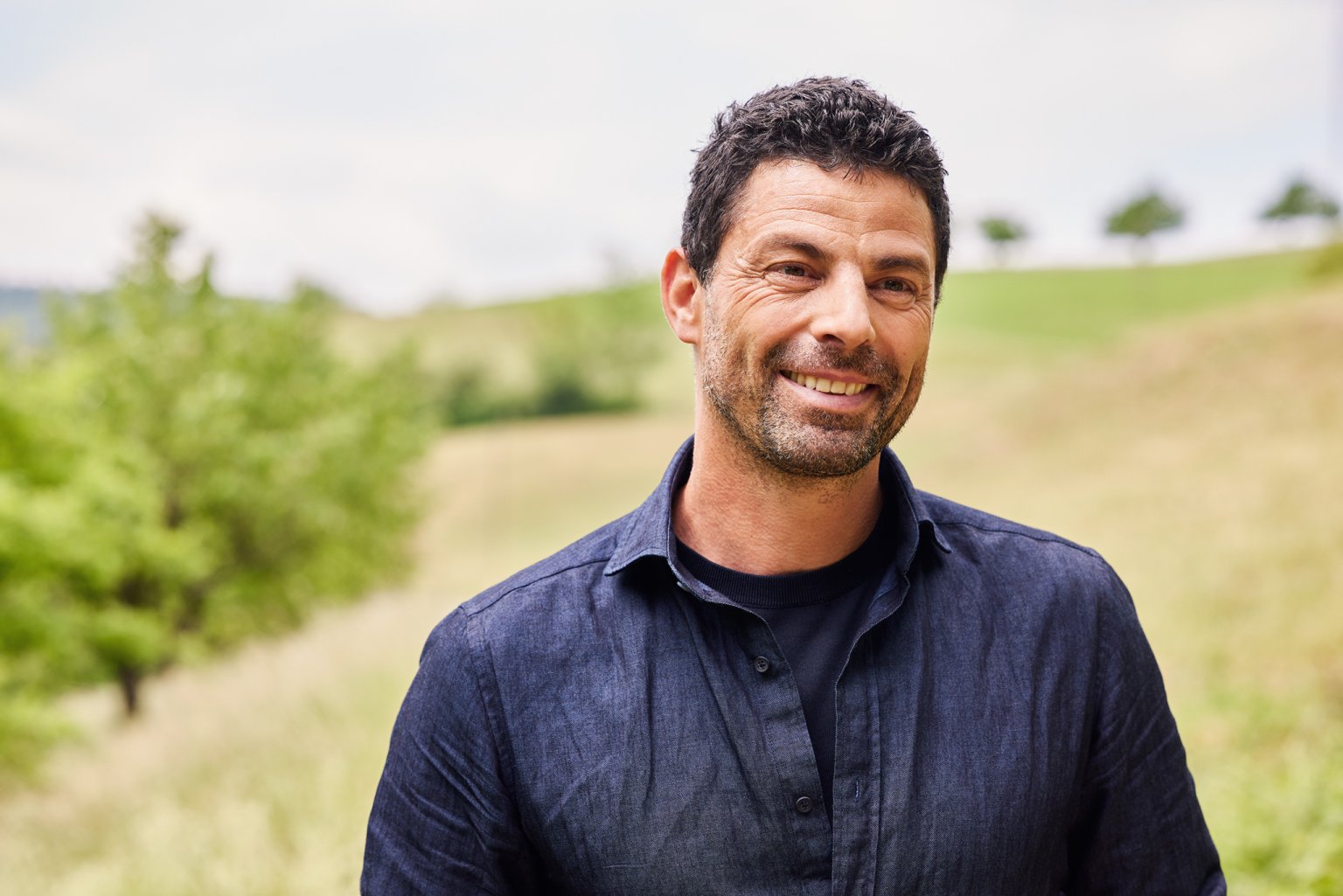 Ein lächelnder Mann vor einer grünen Landschaft.