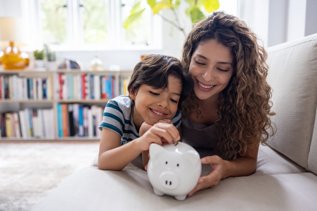 Ein Kind und eine Frau setzen Münzen in eine Sparfigur in einem Wohnzimmer.