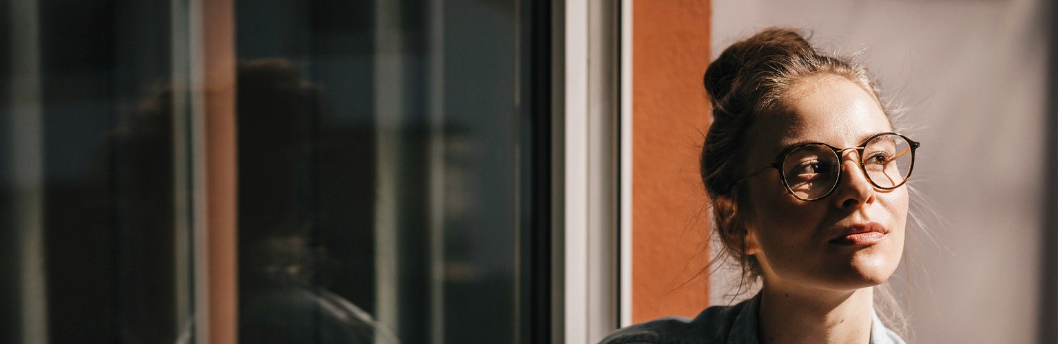 Frau mit Brille, die nachdenklich aus einem Fenster schaut, Sonnenlicht im Hintergrund.