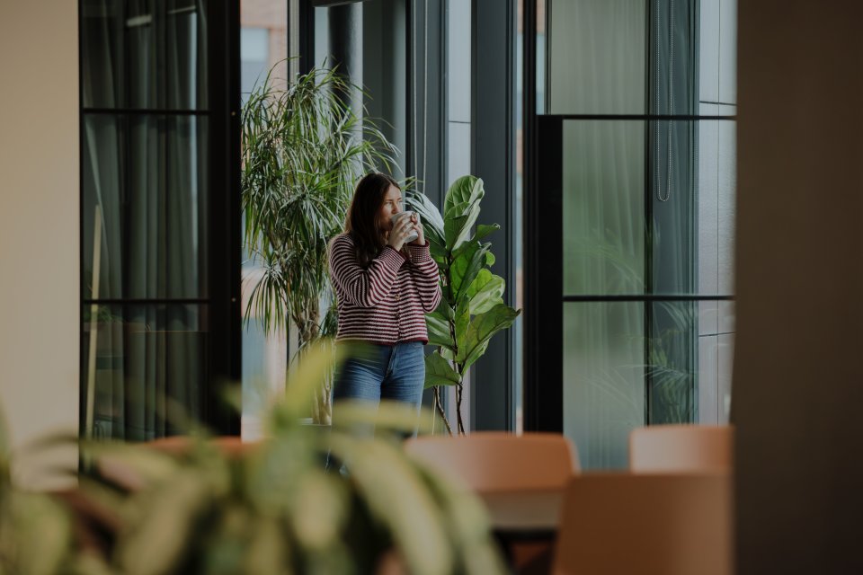 Person sits on armchair with a desk pulled up working on a laptop, two people chat in the background