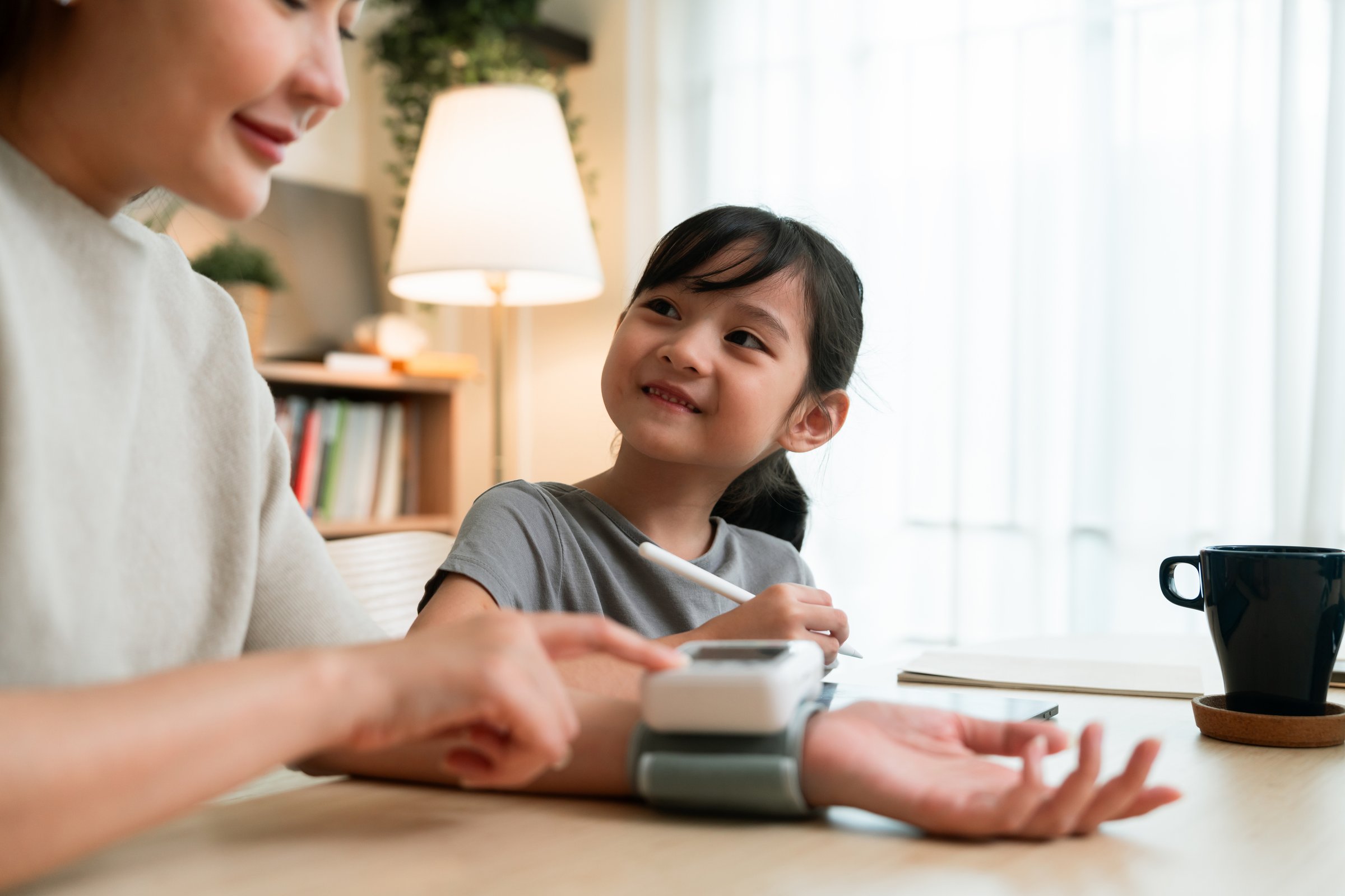 A mother and her young daughter practice using a digital blood pressure monitor at home, enjoying a moment of learning and bonding in a bright, cozy setting.