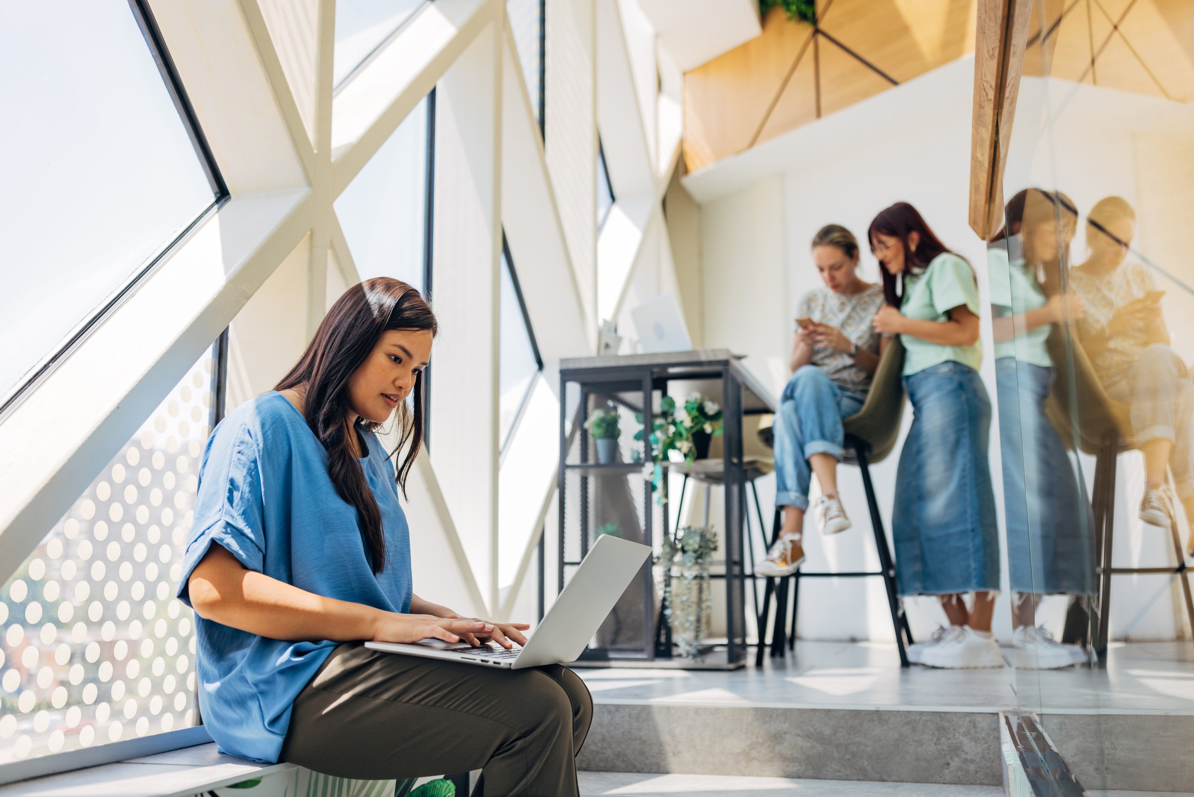 An Asian business woman sits not far from her colleagues in a hybrid business environment and uses a laptop