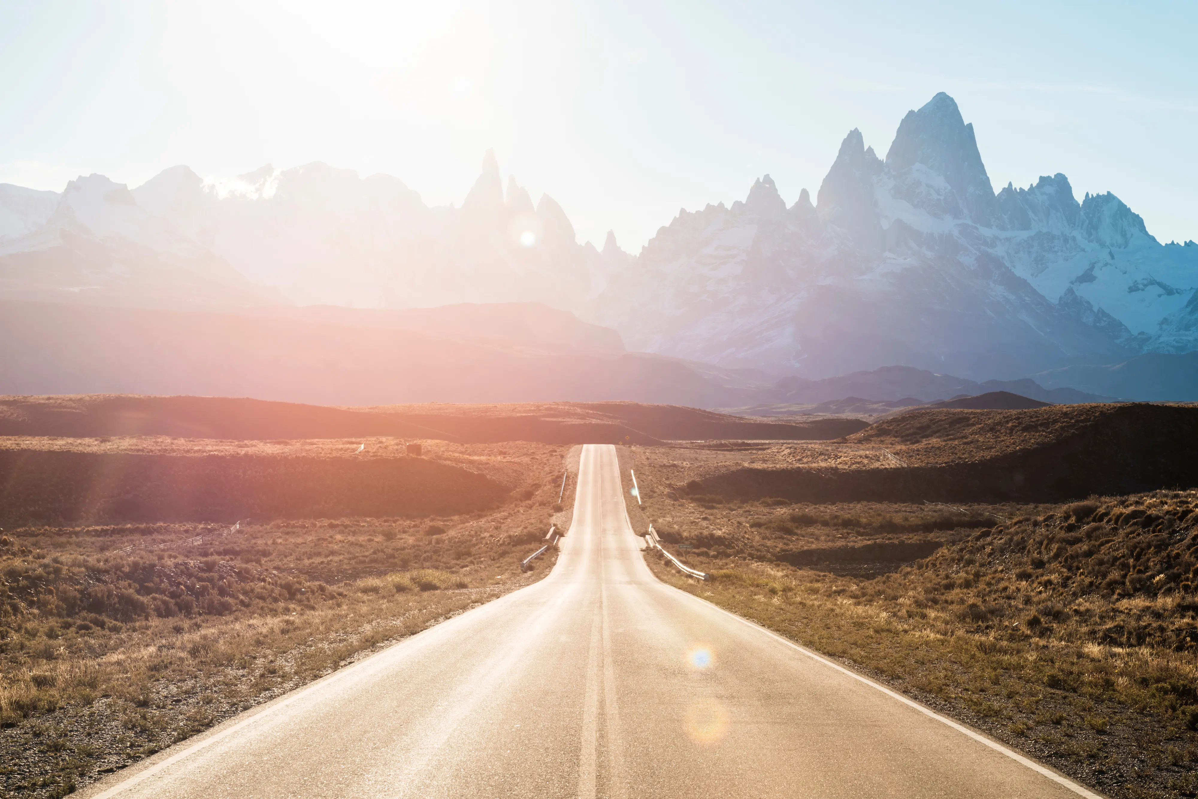 image of a road heading towards mountains with the sun shining