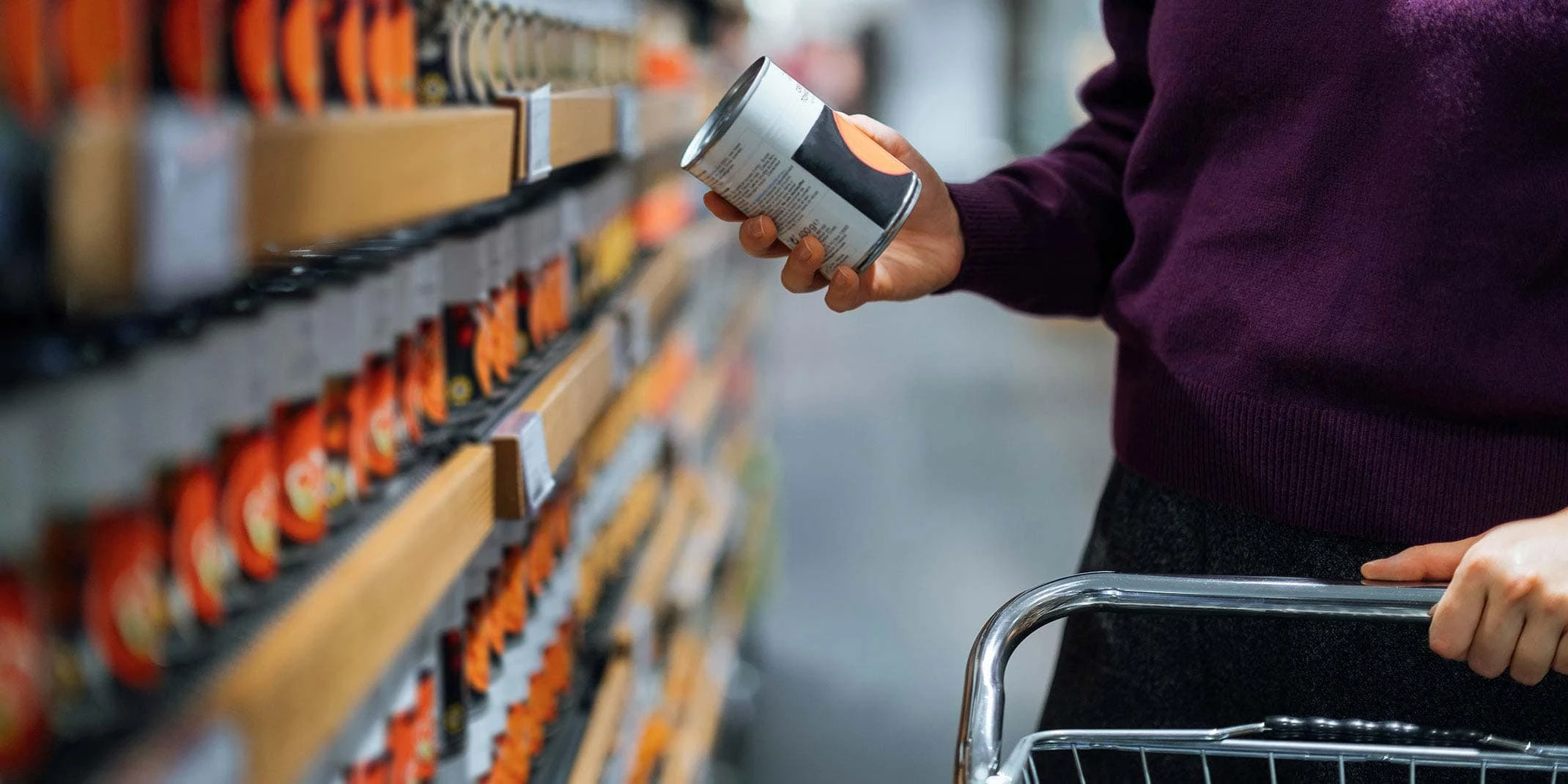 Cropped shot of young woman pushing a shopping cart, grocery shopping in supermarket. She is holding a tin can and reading the nutritional label.