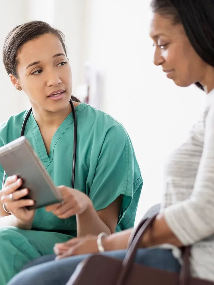Female nurse in green scrubs reviews information on a tablet with a female patient.