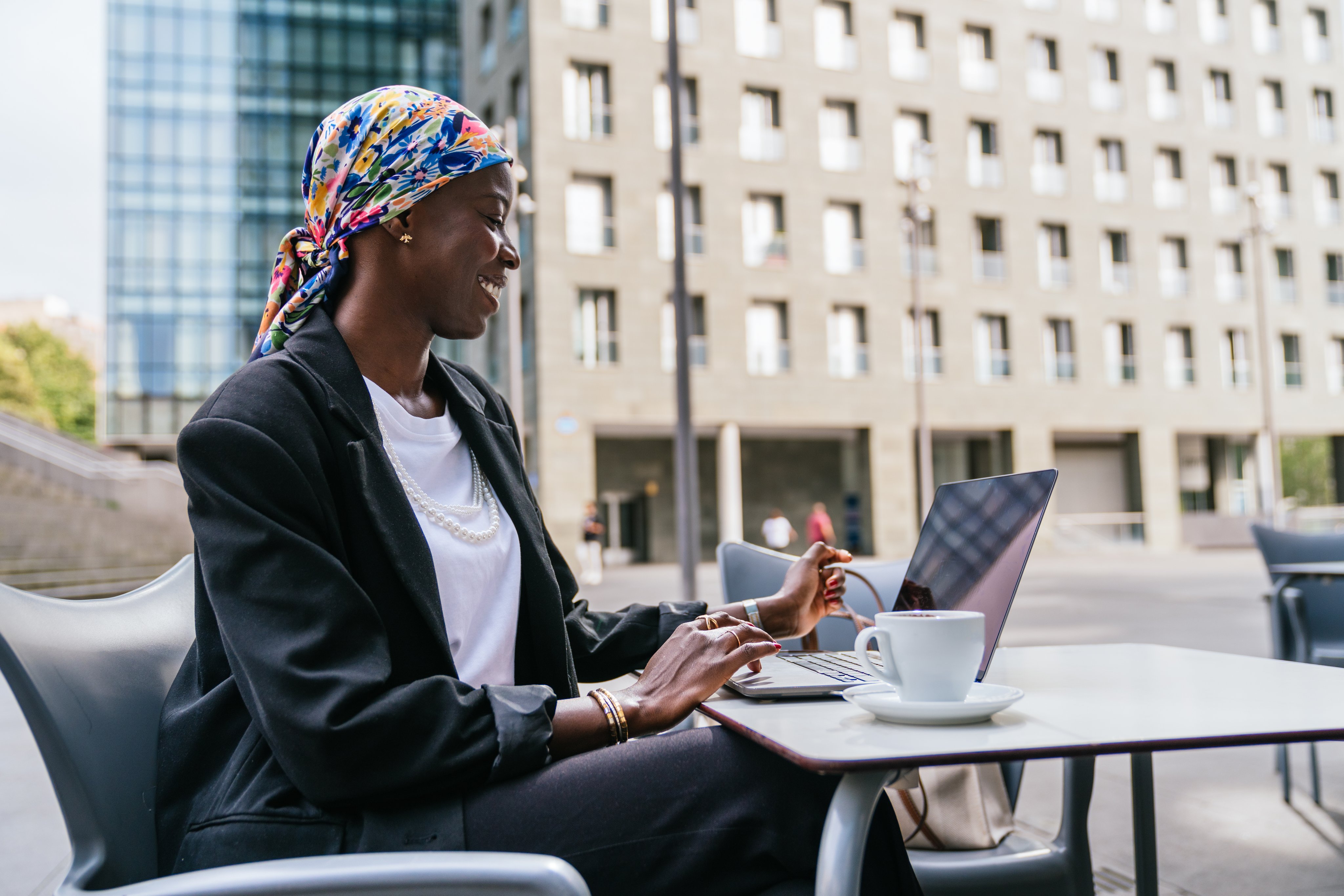 African businesswoman working outdoors on laptop while smiling in a modern urban setting.