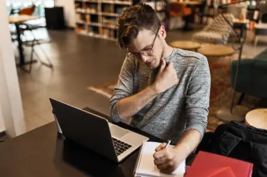 Young man working on his laptop in a library studying writing