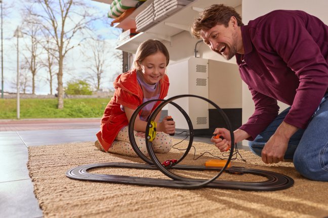 man spelend met kindje met elektrische autobaan op de grond in de garage met een thuisbatterij op de achtergrond
