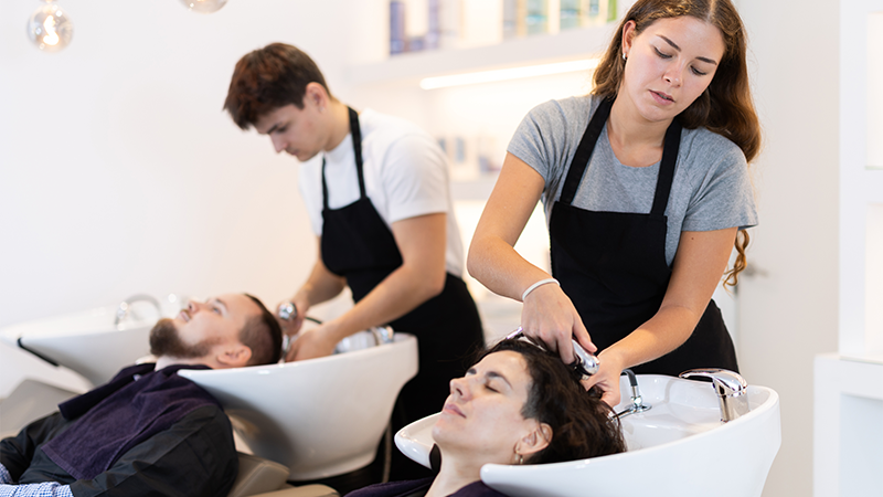 To frisører vasker håret til to kunder som ligger med hodet i vasken.