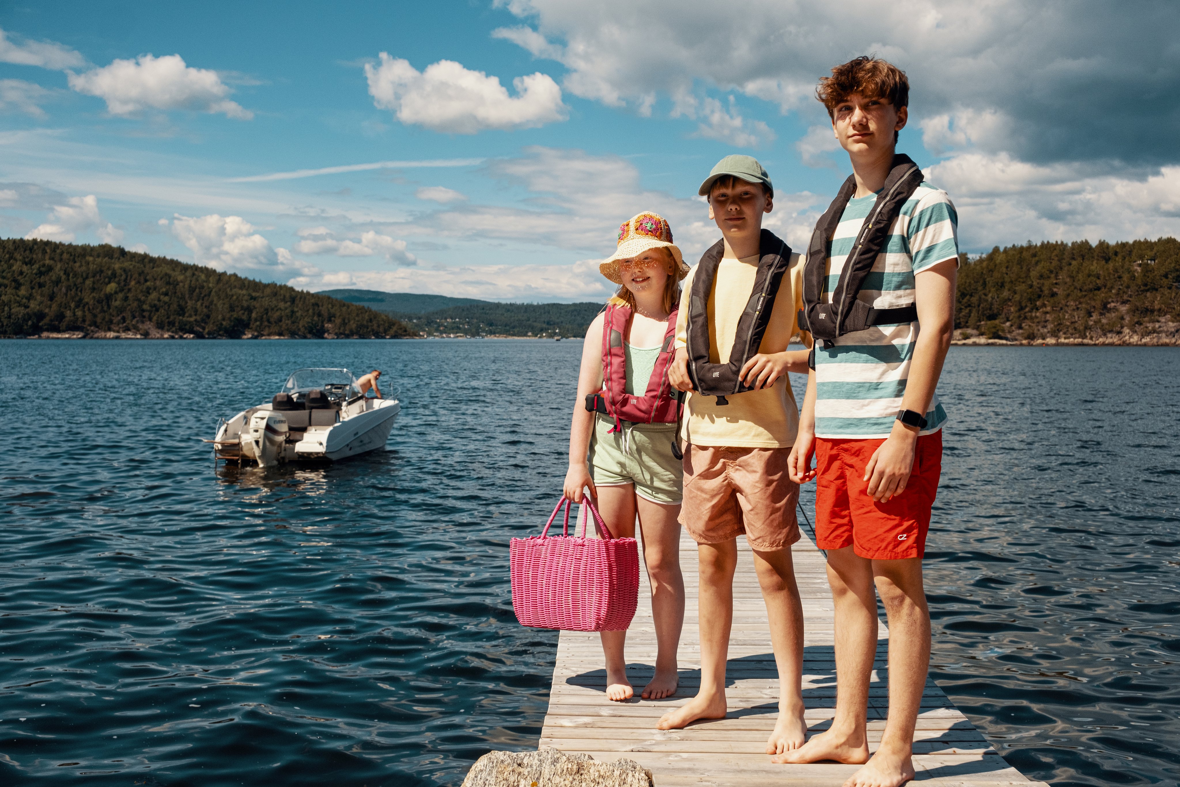 To barn og en ungdom står på en brygge med redningsvester på. Det er sommer, fint vær og en båt kjører i bakgrunnen.