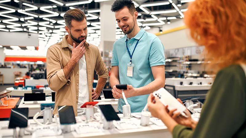 En mann ser på mobiltelefoner ved en butikkdisk mens en ansatt holder en enhet.