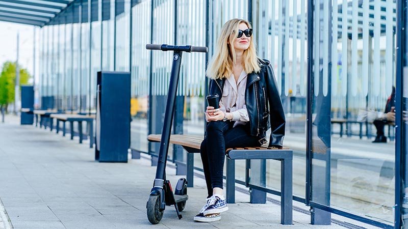 En kvinne sitter og venter på bussen med en elsparkesykkel.