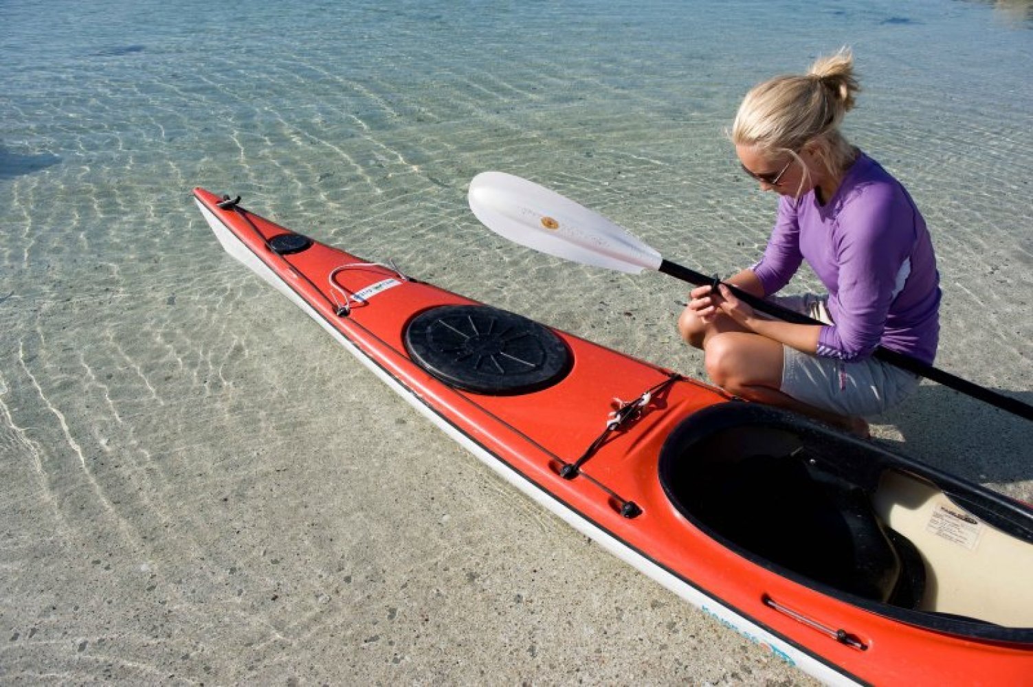 Kvinne sitter på huk på en grunn sandstrand ved siden av en kajakk, mens hun justerer på en padleåre. 