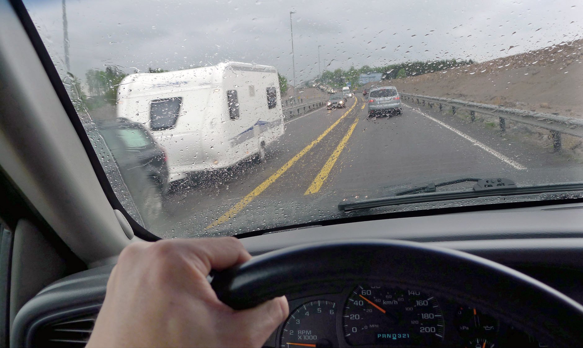 Utsikt fra bilens førersete på en regnvåt motorvei, med en campingvogn og flere biler foran i trafikken