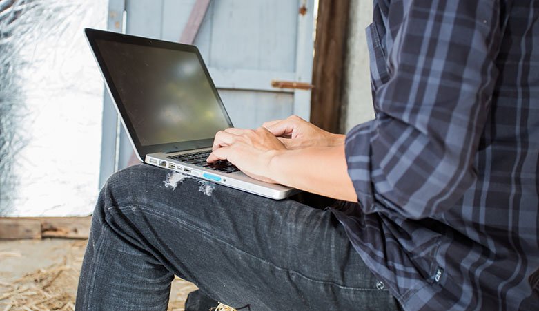 Mann i rutete skjorte og jeans som sitter med laptop på fanger foran en låvedør.