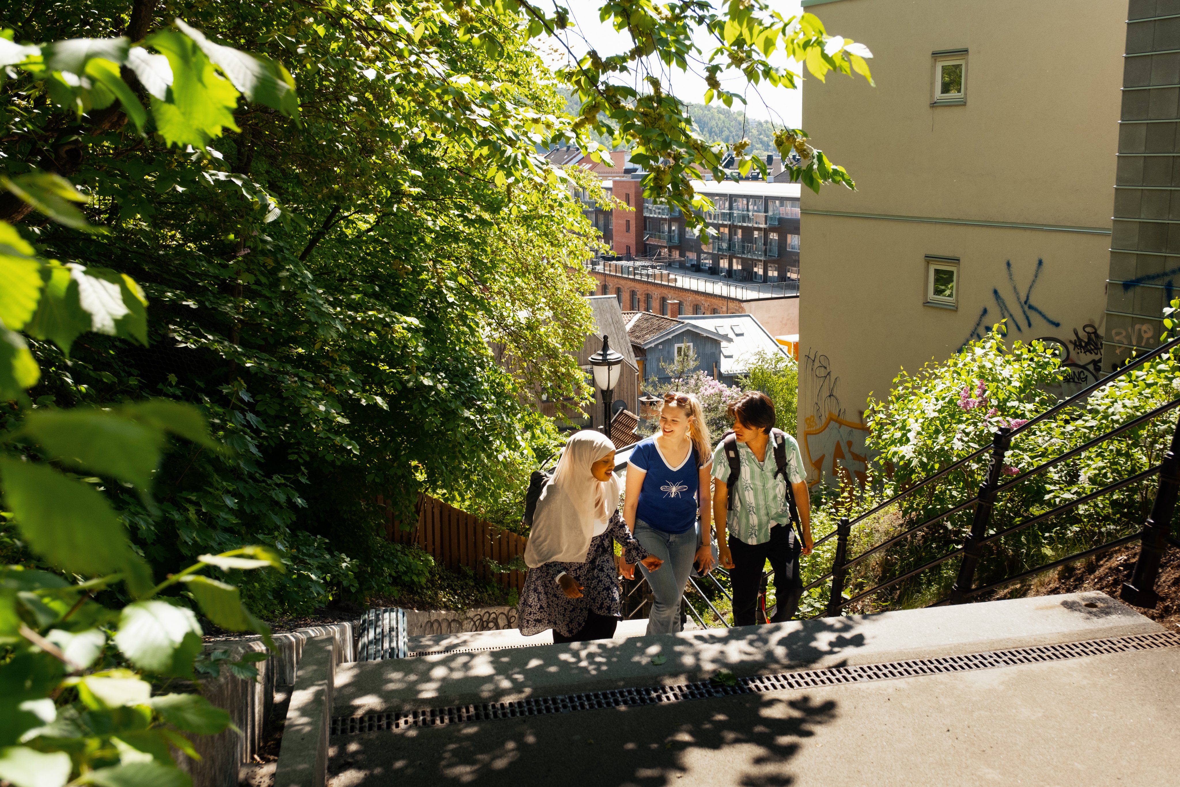 Tre venner går oppover trapp og prater sammen, med bylandskap i bakgrunnen.