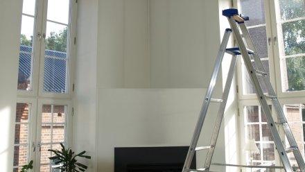 Stue under oppussing med store vinduer og en stige plassert mot veggen. Heksesot er synlig langs veggene.