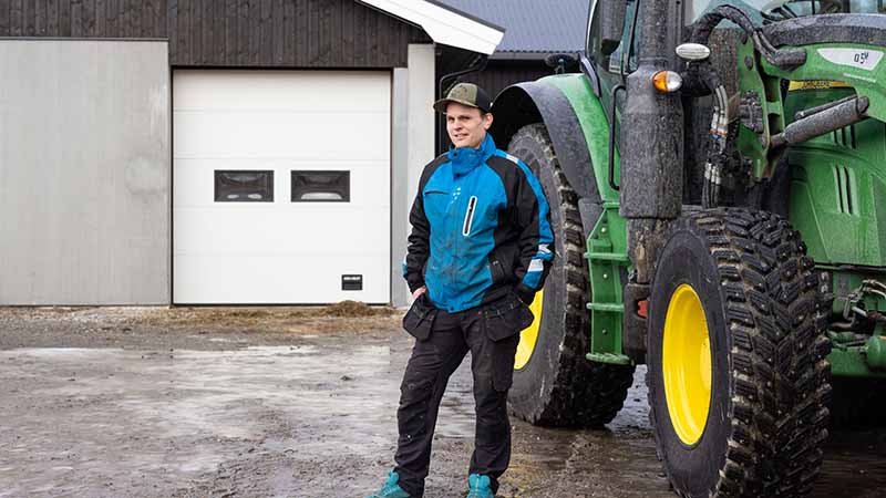Mann i blått arbeidstøy som står med hendene i lommen, med en traktor og grå bygning i bakgrunnen.
