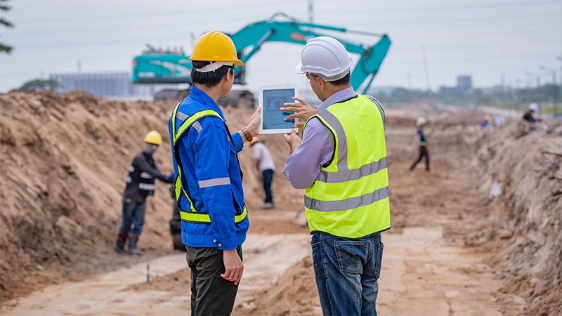 To arbeidere ser på en gravemaskin på et anleggsområde.