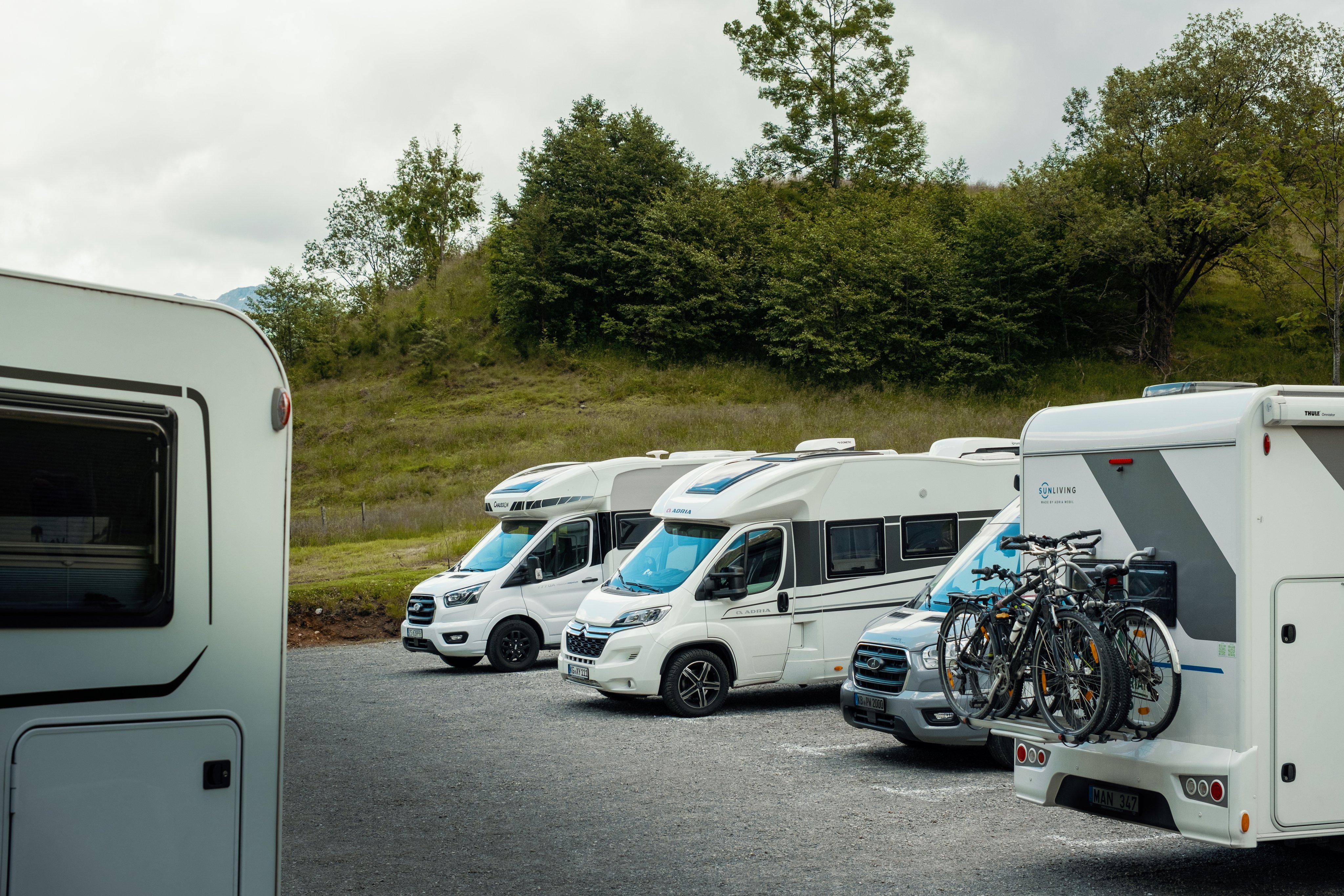 Flere bobiler parkert på en grusplass, omgitt av en åsside og trær.