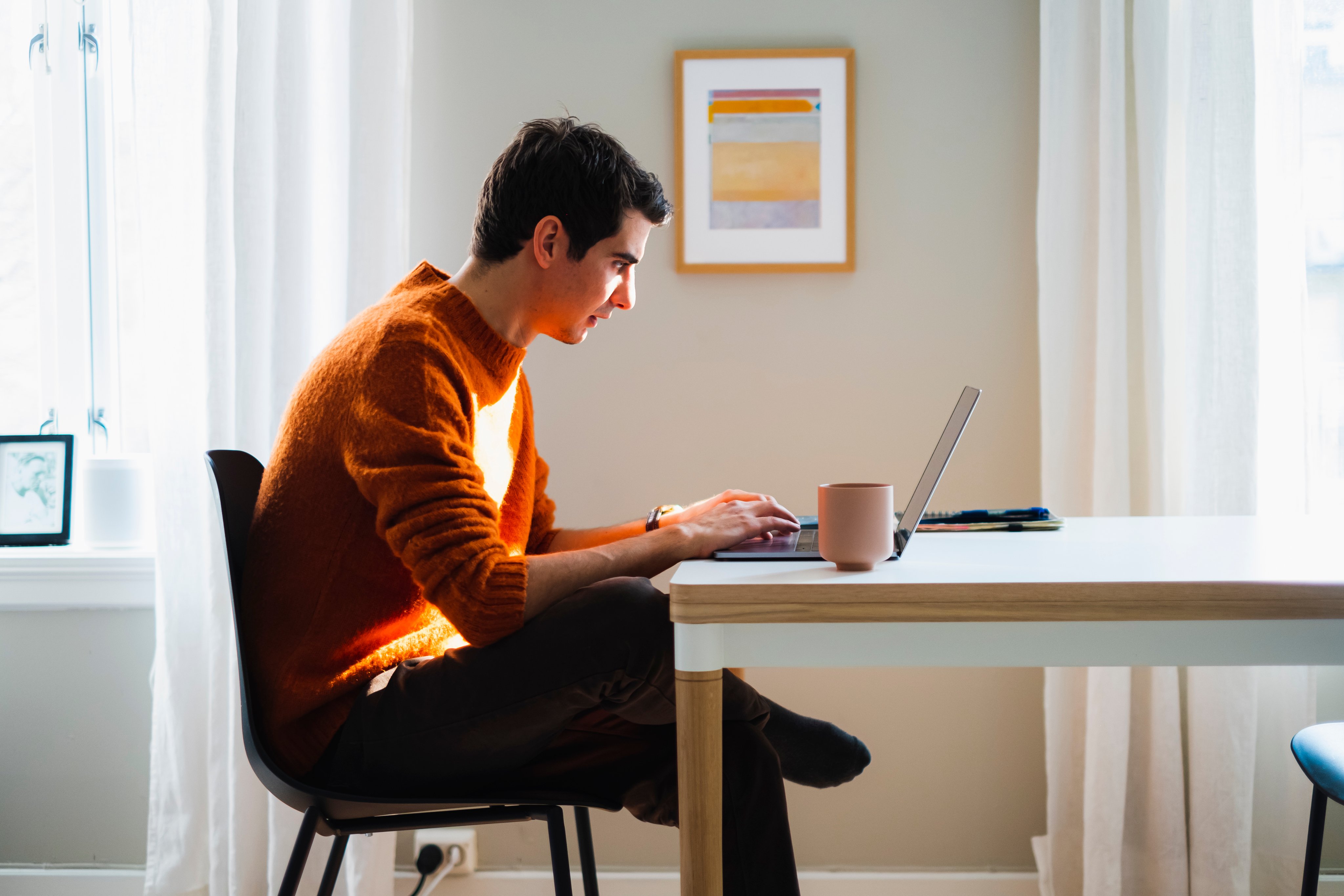 En ung mann sitter i stuen sin og ser på en laptop.